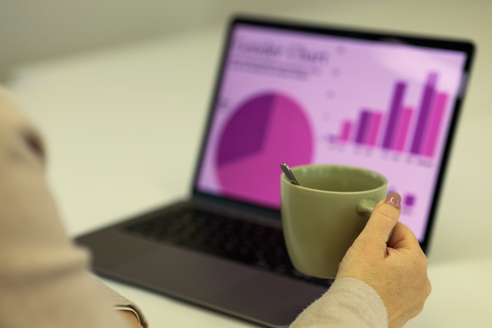 Mão segurando uma xícara de café diante de um notebook com gráficos